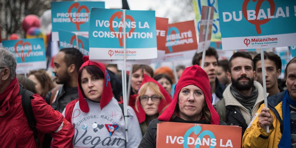Dans la manifestation, des gens portent des pancartes : « Du balai ! », « IVG dans la Constitution », etc.